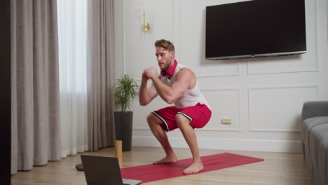 Happy-confident-blond-athletic-man-with-stubble-in-a-white-T-shirt-and-red-shorts-doing-squats-during-his-morning-exercises-while-watching-a-guide-on-his-laptop-in-a-modern-bright-apartment-at-home