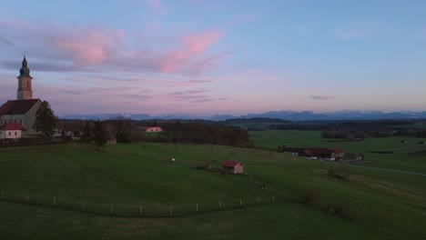 4K-Aerial-view-upon-an-old-Bavarian-catholic-church-chapel-in-the-rural-countryside-by-a-beautiful-cloud-sky-sunset-with-the-alps-mountains-in-the-background-and-green-fields,-farms-and-barns-below