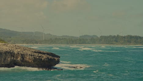 coast of oahu lines the horizon as the white capped turquoise waters of the pacific ocean roll against the boulder lined shore in east honolulu hawaii as wind turbines harvest energy from the wind