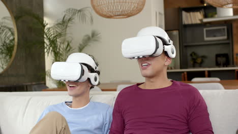 Wearing VR headsets, multiracial gay couple sitting on couch enjoying virtual reality, at home