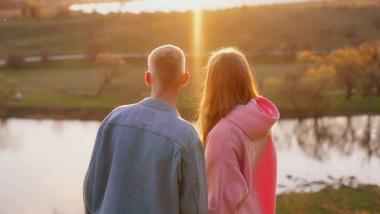 Couple in love in nature at sunset. Beautiful young girl and her boy friend admiring the setting sun over the river in autumn.
