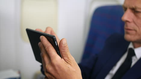 primer plano de un hombre de negocios maduro que usa un dispositivo de tableta en un avión que trabaja en la cabina de un avión de pasajeros