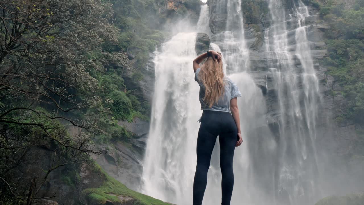 A woman stands in awe before the roaring Bomburu Ella Waterfalls, surrounded by mist, jungle, and rocky cliffs in the lush highlands of Nuwara Eliya, Sri Lanka.