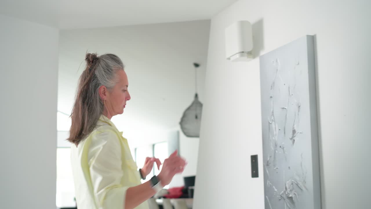 After picking up canvas, woman is carrying it into hallway, lifting and aligning to hang