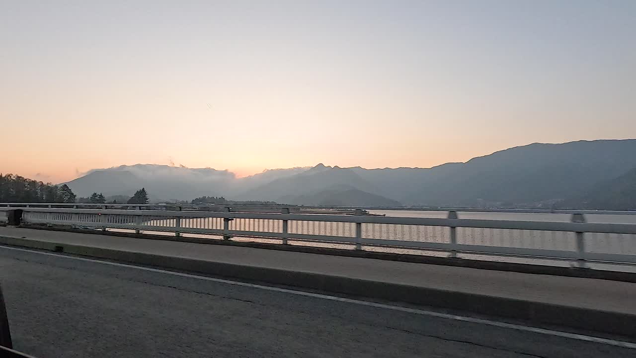 Vehicles cross a bridge over a serene mountain lake at sunrise, with a tranquil atmosphere and soft lighting