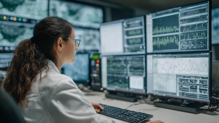 A focused professional in a lab coat analyzes complex data on multiple computer screens, showcasing advanced technology in a high-tech monitoring environment