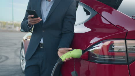 successful businessman charging electric car on station and using modern smartphone for work african