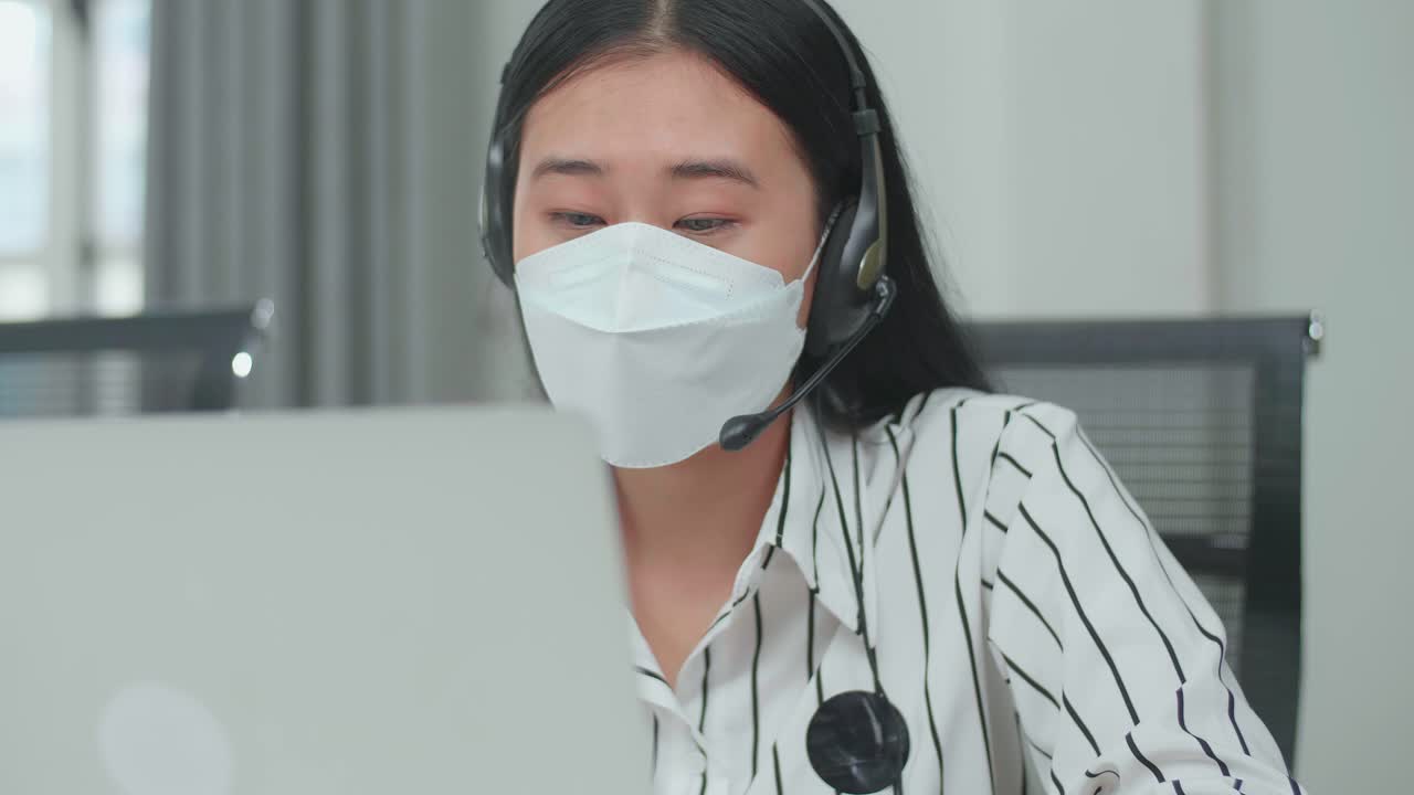Close Up Of An Asian Woman Call Center Agent Wearing Headset And Mask Typing On The Computer While Speaking To A Customer On The Call During Working With Colleagues At The Office