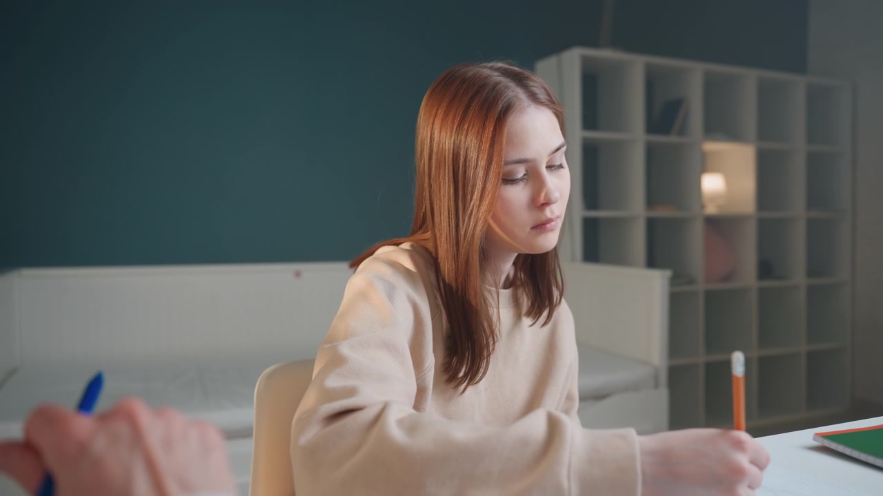 Un adolescente bianco è concentrato sui compiti scritti, mentre il tutor a domicilio osserva dallo sfondo. La matita scorre sulla pagina, segno di silenziosa concentrazione e determinazione. L'insegnante di grammatica offre una guida discreta. Scrivania ordinata, lampada.