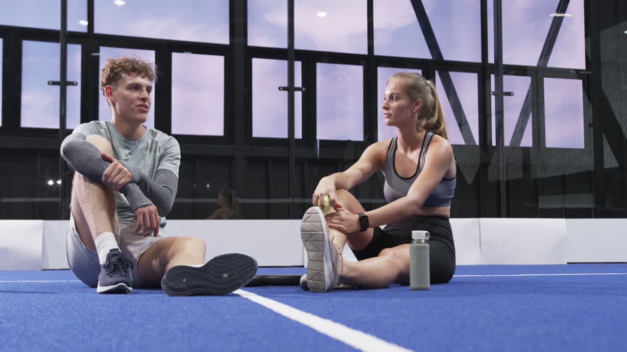 Young man and woman resting on padel indoor court, holding tennis ball, enjoying conversation