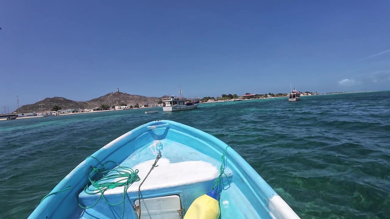 POV da una piccola imbarcazione che si avvicina al villaggio di Gran Roque attraverso le acque turchesi dei Caraibi, con montagne costiere e barche in lontananza
