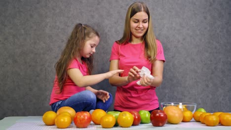 mother and daughter working together in the kitchen. healthy organic fruit on the table. beautiful woman and little girl peeling fruits.