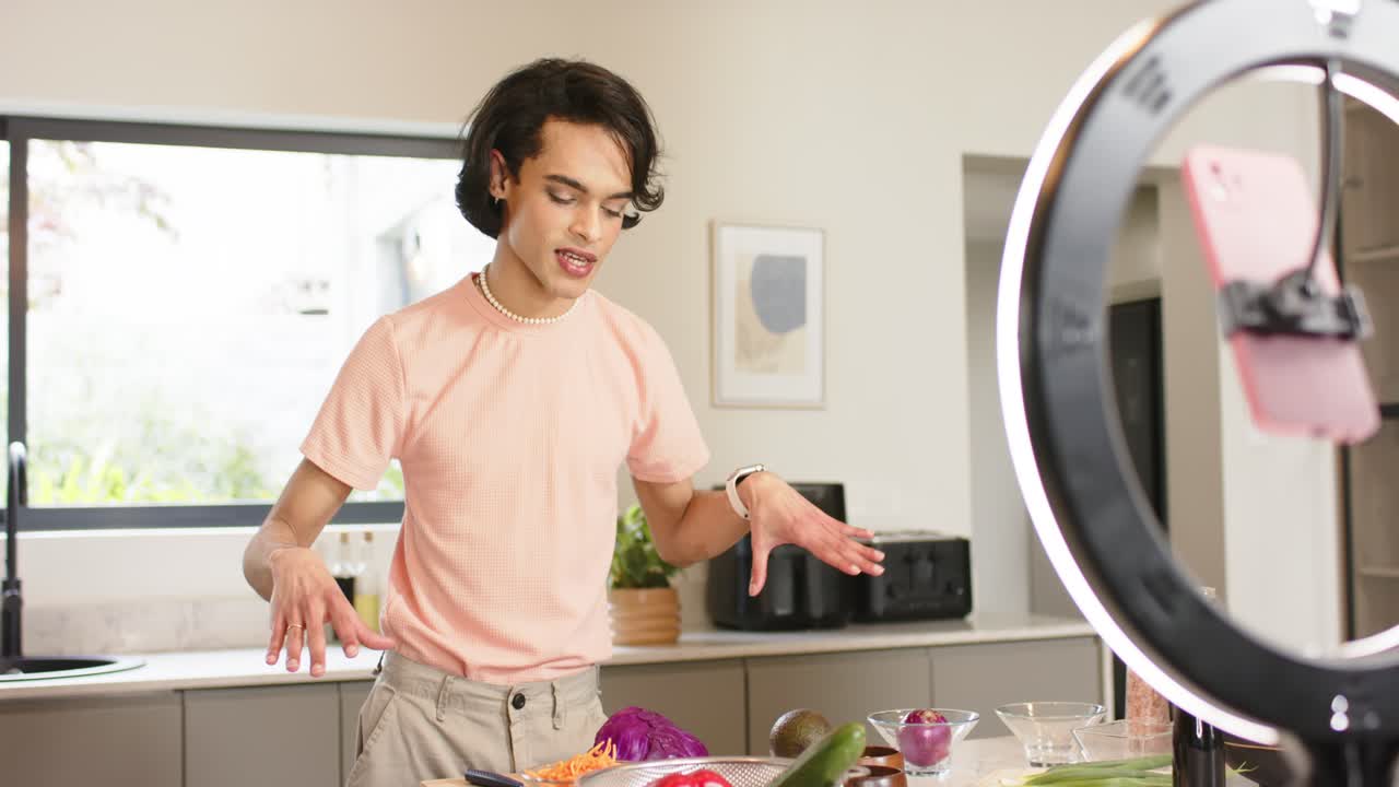 Uomo che saluta gli spettatori e condivide la ricetta di un'insalata sull'isola della cucina mentre mescola le verdure illuminato da una ring light