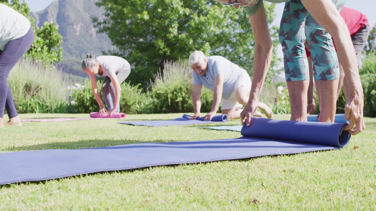 남녀 노인들의 다양한 그룹이 정원에서 운동한 후 요가 매트를 굴러 올리고, 느린 동작