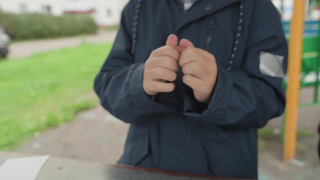 屋外で小さな工作を準備する白人の小学生。遊び場のベンチで指先と紺色のジャケットに集中している。小さな部品を触覚で操作する様子をクローズアップで捉えた連続写真、集中した表情、作業の様子。