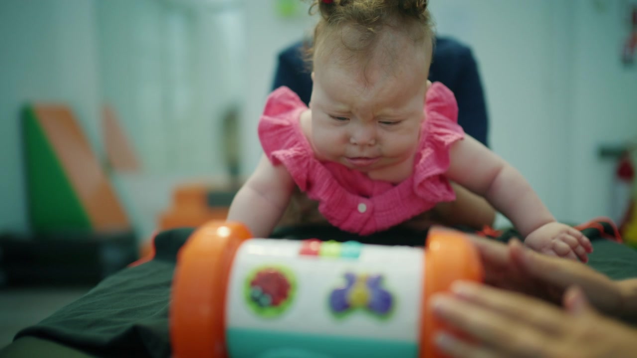 Bebê pequeno interagindo com um brinquedo colorido, recebendo apoio de intervenção precoce e fisioterapia de um profissional de saúde.