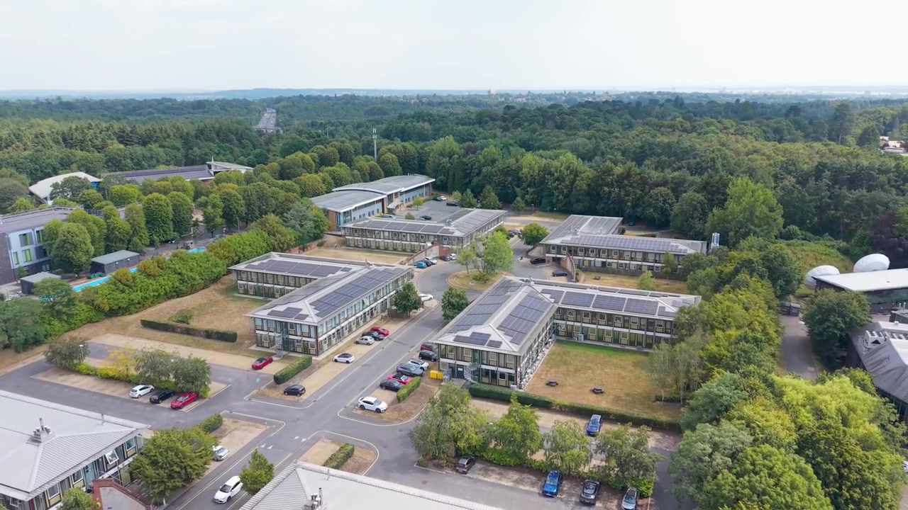 Drone reverses away from Southampton Science Park, revealing solar panelled rooftops of office buildings surrounded by dense forest, England.
