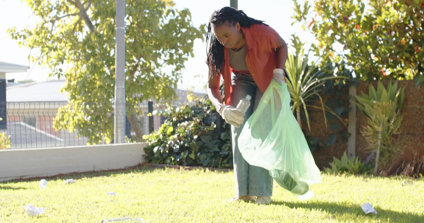 African woman holding green bag with white gloves, bending, picking bottles in small yard to clean
