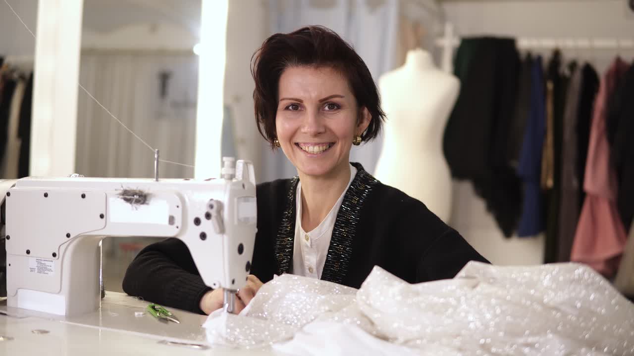 Portrait of female tailor working on sewing machine. Caucasian woman using a sewing machine at studio or in workshop. Woman cheerfully smiling to camera and back to the process