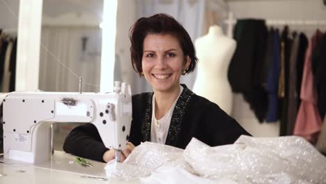 porträt einer weiblichen schneiderin, die an einer nähmaschine arbeitet. kaukasische frau mit einer nähmaschine im studio oder in der werkstatt. frau lächelt fröhlich zur kamera und zurück zum prozess