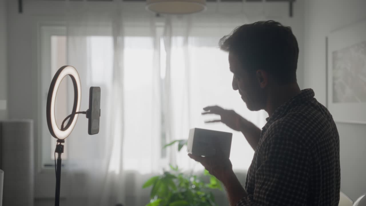 Una celebrità dei social media o un* vlogger influencer sta girando un video di unboxing per la creazione di contenuti online. In piedi nel suo appartamento di fronte a un supporto per telefono con ring light, apre una scatola e recensisce il contenuto.