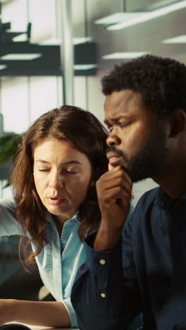 Vertical video Employee and coworker looking over revenue figures on PC in office