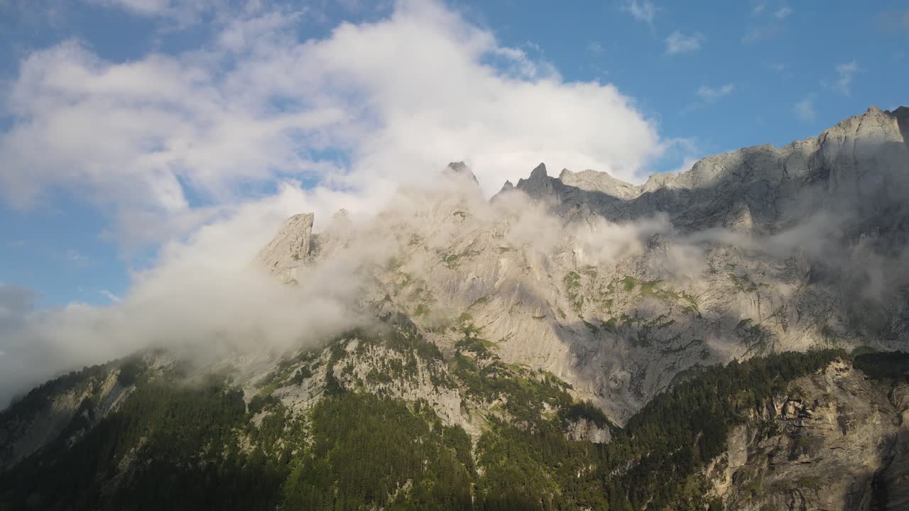 Nubi basse si spostano lentamente su una frastagliata catena montuosa alpina lungo la Via Alpina in Svizzera. La luce naturale rivela ripide pareti rocciose, pendii boschivi e cambiamenti climatici sulle alte Alpi.