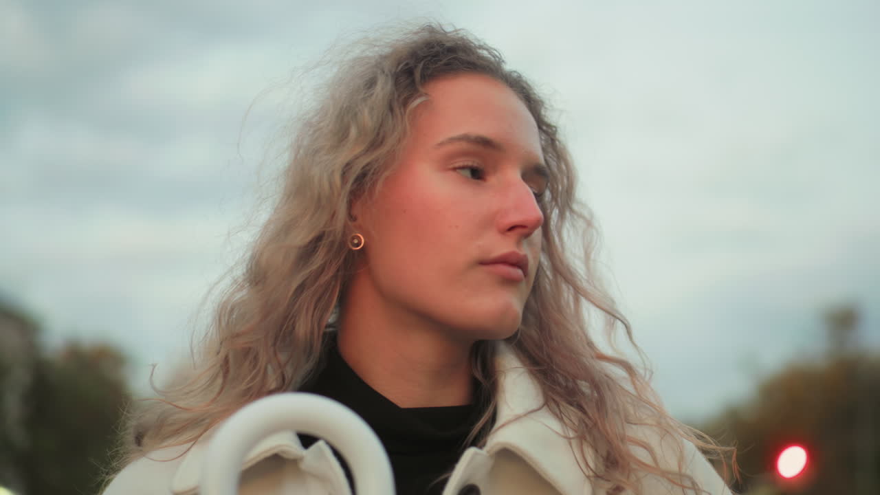 Calm woman in white coat walking through urban setting holding umbrella closely with thoughtful expression under cloudy sky in peaceful moment of solitude during soft light of evening