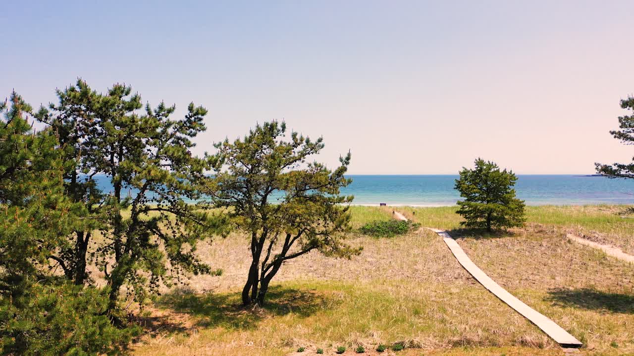 Aerial drone video flying over a boardwalk bridge winding through tall green grass, leading toward the peaceful ocean and sandy beach in Saco, Maine. Scenic coastal walk with waves and blue sky.