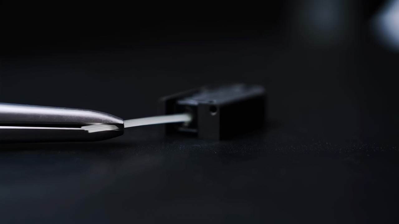 Close-up macro view of metal tweezers carefully placing a thin white strip into a small black mechanical component against a dark background with soft