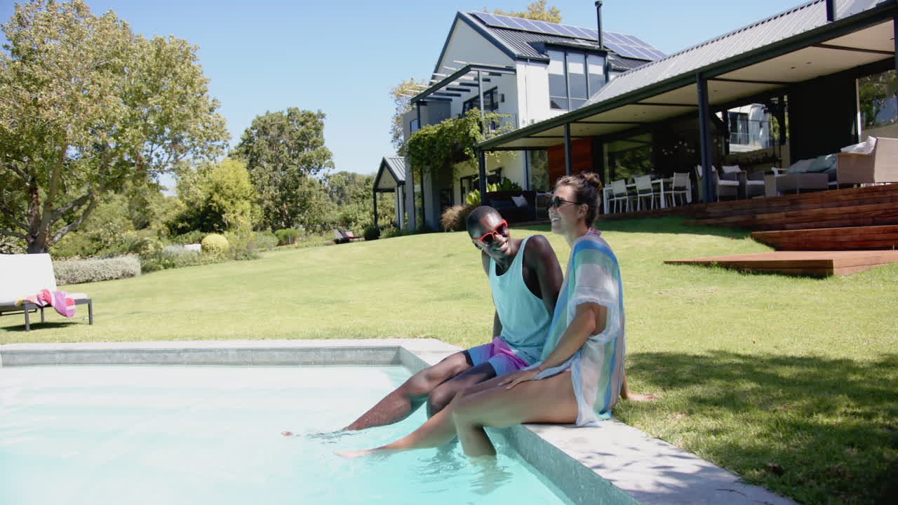 Relaxing by pool, diverse couple enjoying sunny day at modern house with garden, copy space