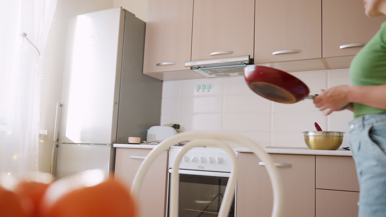 Close up of oranges in foreground with woman in green shirt holding frying pan in kitchen, preparing to cook near modern cabinets and appliances in bright interior
