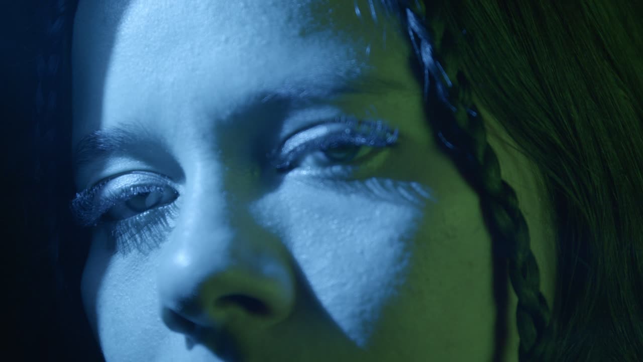 Dramatic close-up shot of a woman's intense gaze styled with bold lighting. Intricate braid captured under striking blue and green hues create a mysterious, contemplative mood