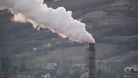 thick smoke billows from a large industrial chimney, dominating the skyline above a small village nestled in a hilly landscape