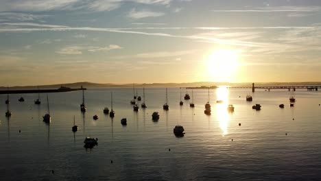 Boats-silhouetted-on-golden-shimmering-harbour-ocean-surface-aerial-view-slowly-tilting