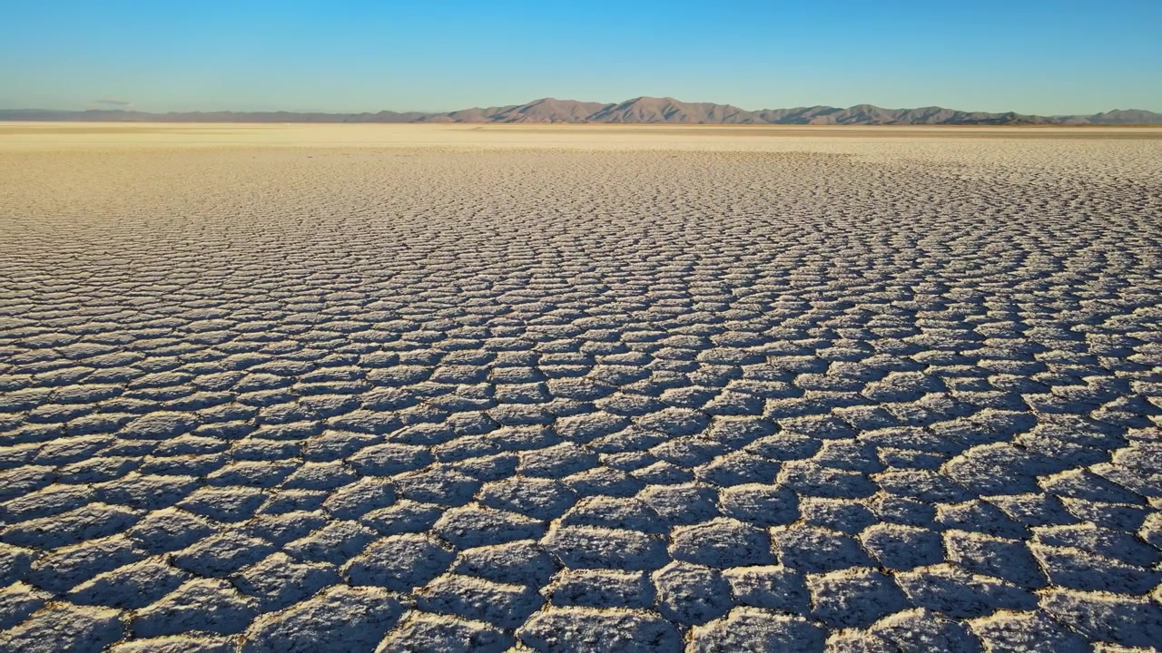 Salt polygons stretch across the wide Salinas Grandes salt flat with distant mountains under clear blue sky, useful for themes of drought, climate change, isolation and abstract nature.