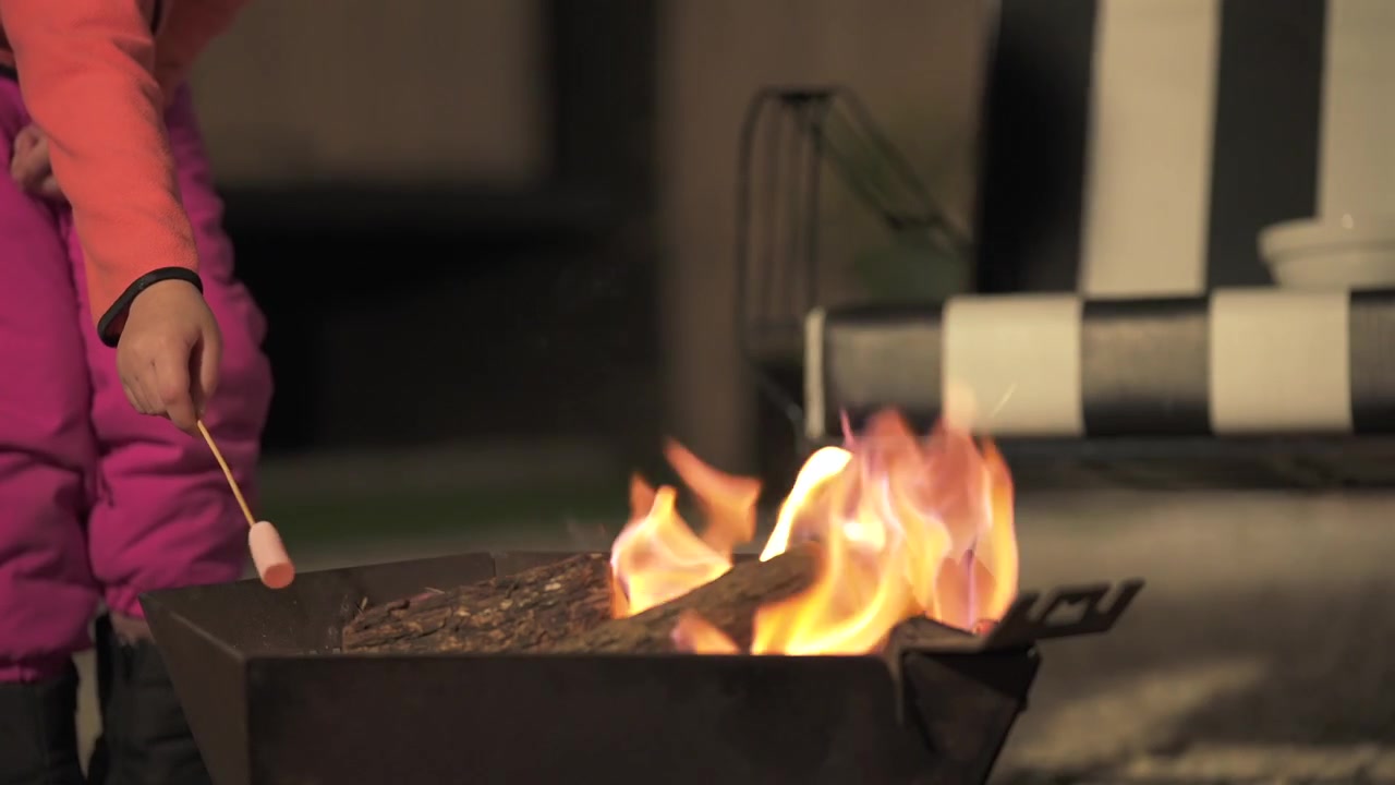 Ragazza con una giacca rosa che tiene un bastoncino di legno e arrostisce un marshmallow vicino alle fiamme di un braciere da esterno. Vista ravvicinata statica.