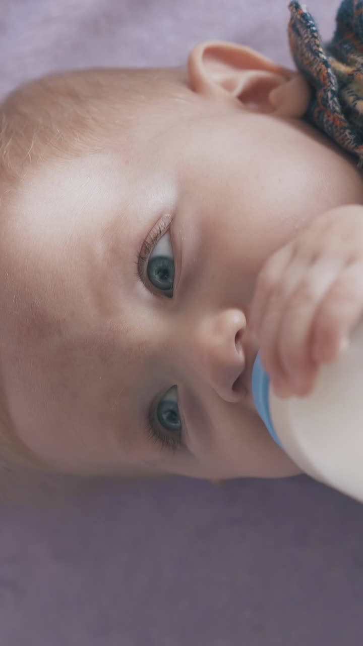 cute little boy eats delicious milk mix from bottle lying on soft bed with pink plaid in light room close view