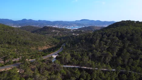 road winding through a vibrant green forest in ibiza, spain, leading down towards the beautiful san antonio bay. fabulous aerial view flight overflight flyover fly push forward drone