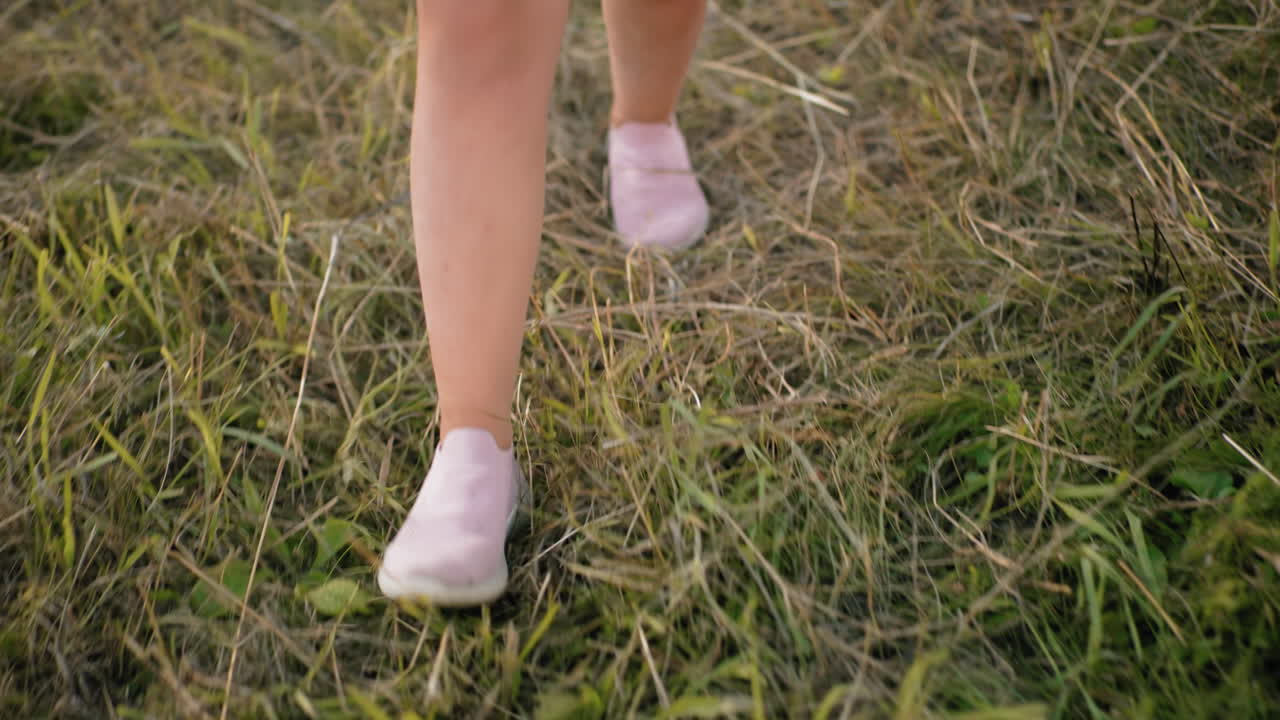 건조한 풀을 고 있는 사람이 분홍색 슬립 스니커를 입고 있는 클로즈업, 초록색과 노란색 잔디가 인 불평등한 지형에서 자연스럽게 걸어가는 단계