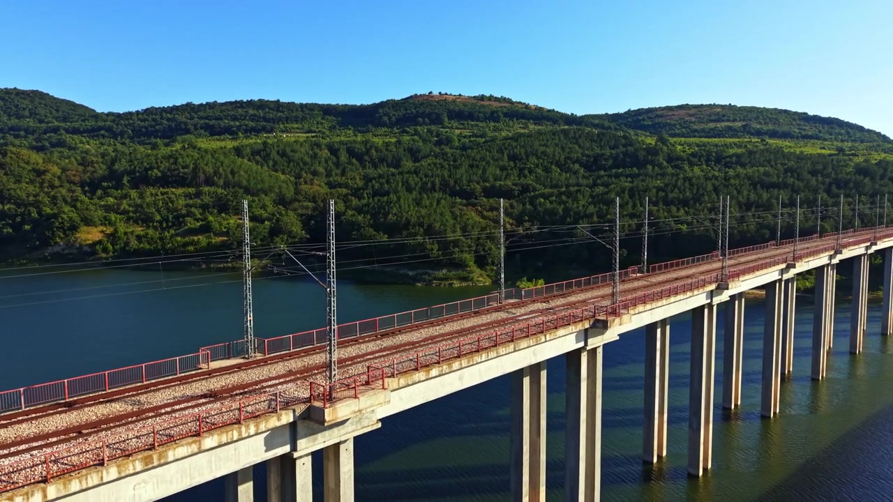 Vista de uma ponte ferroviária sobre um rio com colinas verdes ao fundo sob luz solar intensa