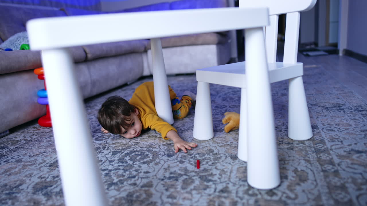 Small kid lies on the floor near his desk. Toddler tries to get the cap from a felt pen. Boy stands up and walks to the seat.