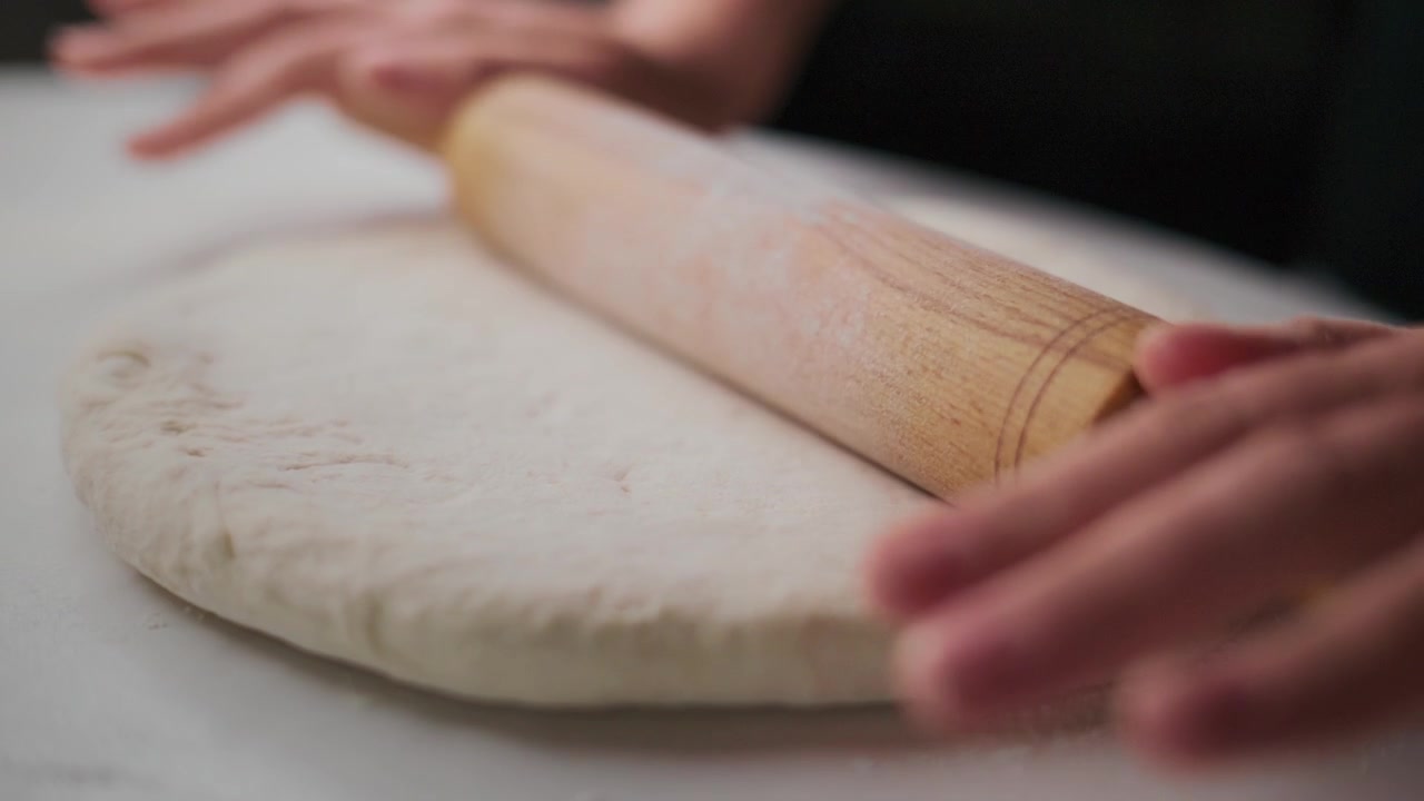 Una persona stende l'impasto con un mattarello di legno su una superficie bianca. Le mani applicano una leggera pressione mentre il mattarello appiattisce l'impasto. L'attenzione rimane sul movimento e sulla consistenza dell'impasto.