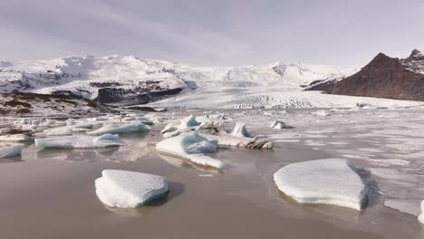 iceland’s fjallsárlón glacier lagoon - drone slowly pulling back over icebergs and frozen water with snow-covered mountains and glacial terrain.