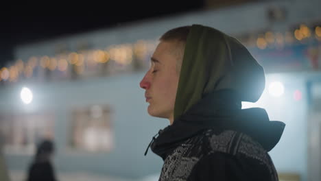 close up side view of young man wearing green hoodie and winter jacket walking slowly outdoors in cold weather during night, soft festive lights and green lens flare visible in background
