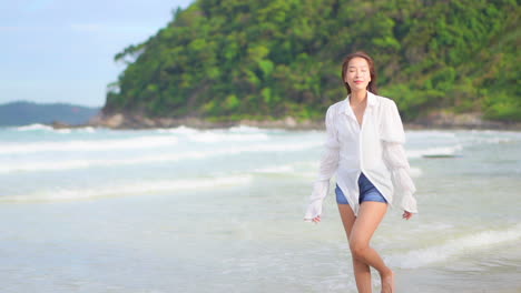 Asiatische-Frau,-Die-Einen-Spaziergang-Am-Meer-Entlang-Macht-Und-Blaue-Shorts-Und-Ein-Weißes-Hemd-Mit-Unscharfen-Wellen-Trägt,-Die-Das-Ufer-Im-Hintergrund-Einer-Tropischen-Insel-Erreichen