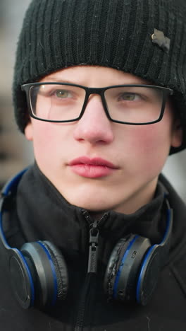 close-up of a young boy wearing a black outfit and beanie, looking focused and contemplative, showing his glasses and a pair of headphones resting around his neck