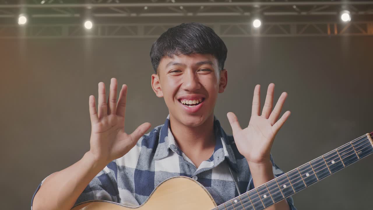 기타를 들고 있는 아시아인 십대가 손바닥을 보여주고, 와우라고 말하고, 무대에서 미소 짓고 있습니다.