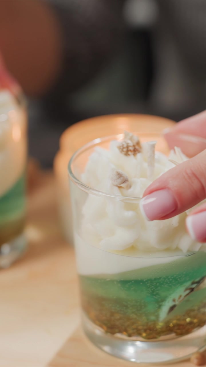 Close up of hand with manicured nails gently touching layered candles in glass featuring white whipped cream and decorative elements. scene includes soft lighting on wooden surfaces