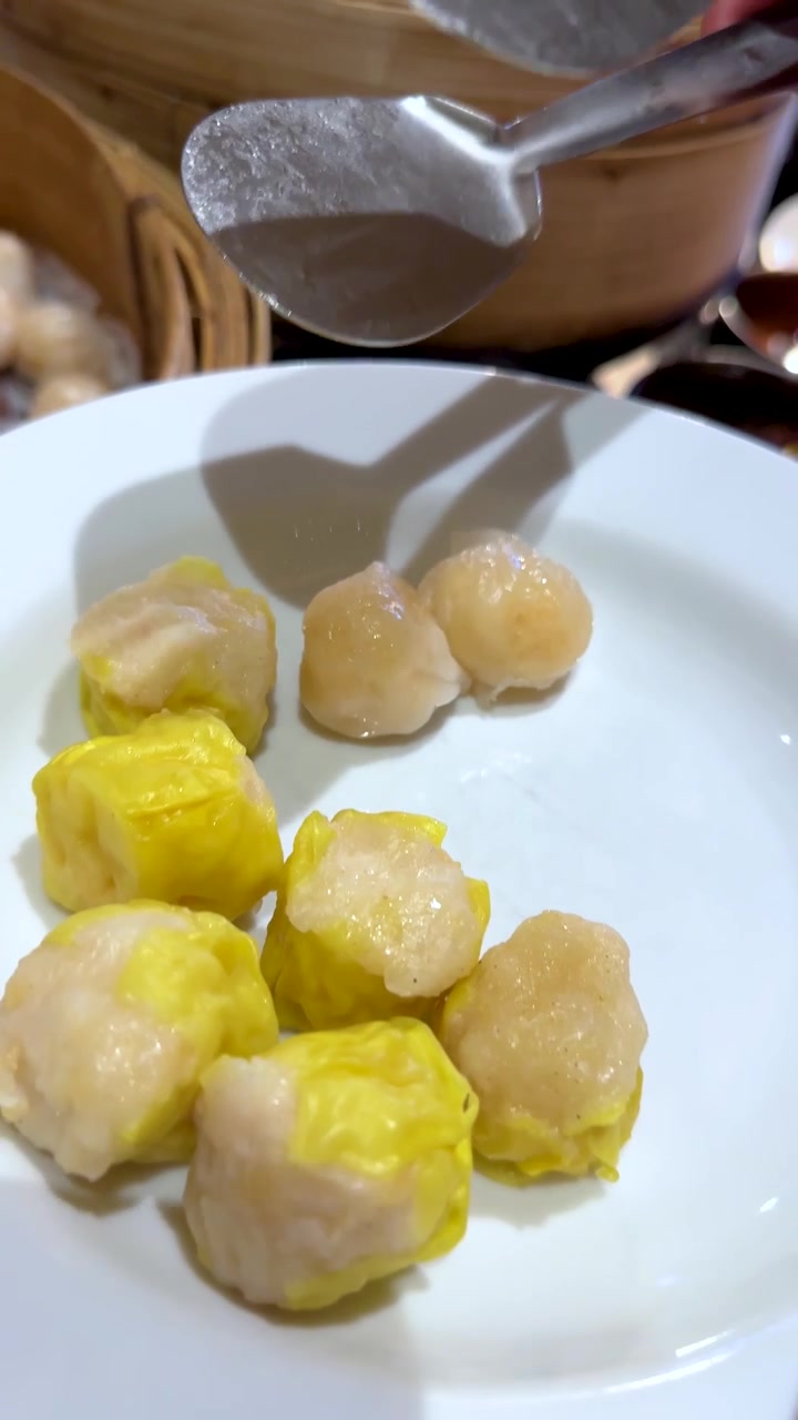 Penjepit logam mengambil siomay kuning dan pangsit udang dari kukusan bambu ke piring.